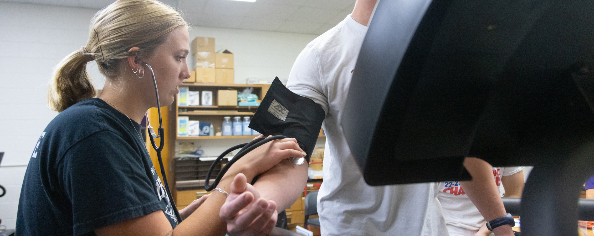 A student uses a stethescope to check the heart rate with of another who is on a treadmill.