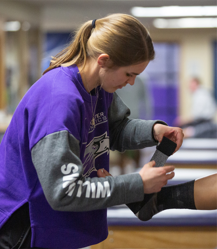 A person in a Warhawk Athletics shirt tapes the ankle of a person off camera.