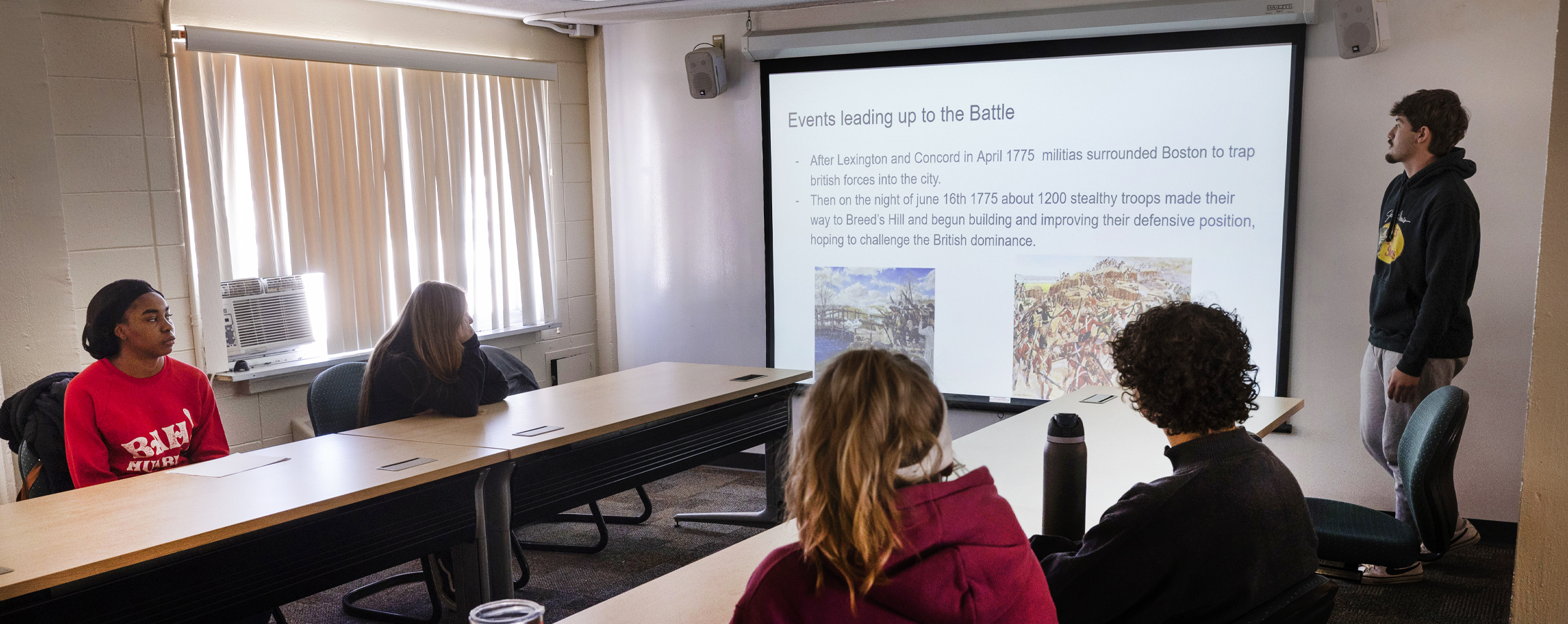 Four students sit at long tables in a classroom and view a presentation.