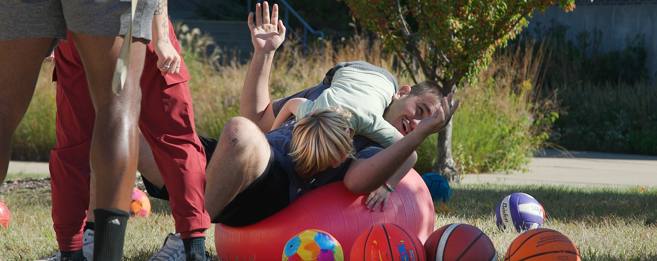 A student lays on a large red exercise ball and laughs as a child jumps on him.