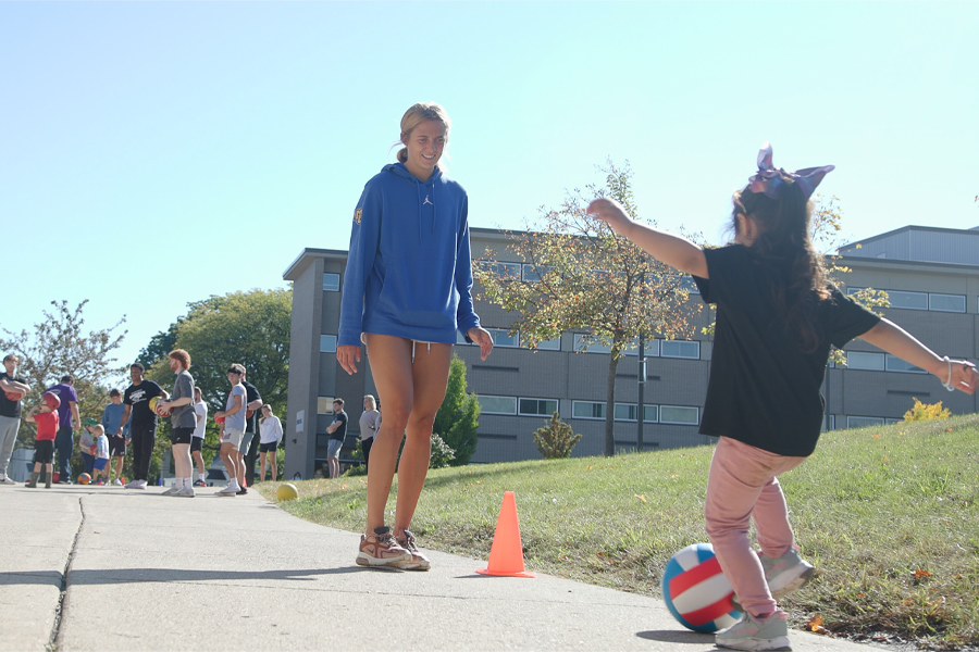 A student smiles as she watches a child dribble a soccer ball through orange cones.