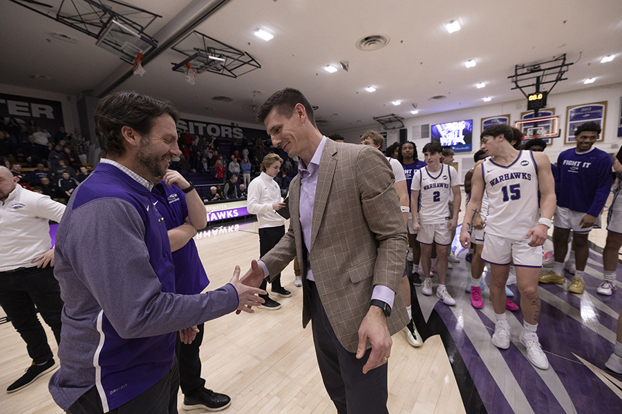 An athletic director in a purple vest shakes hands with a basketball coach on a basketball court with basketball players in the background.