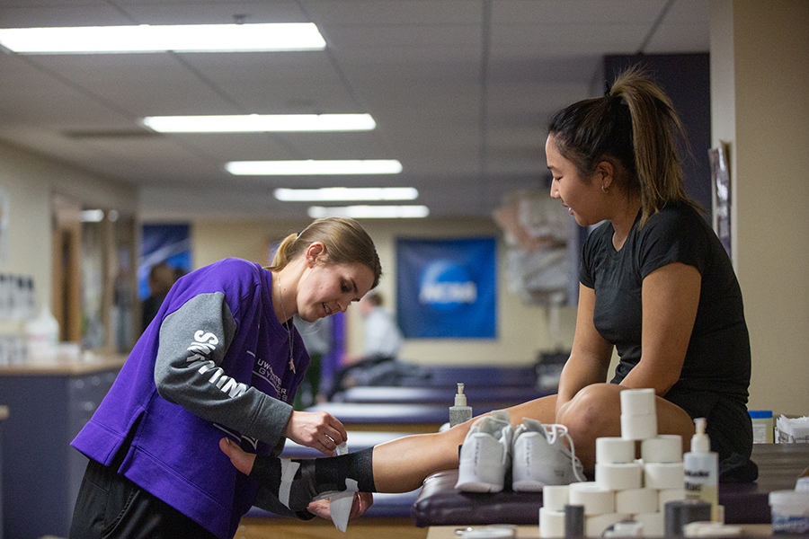 A person in a purple Warhawks shirt tapes the ankle of another person.