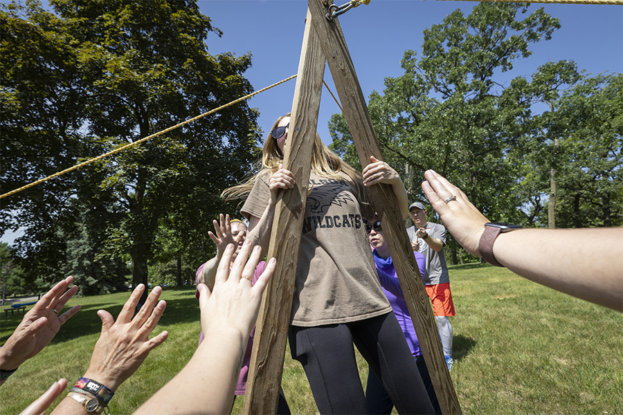 A group of people outdoors work together to form an A frame with wooden posts.