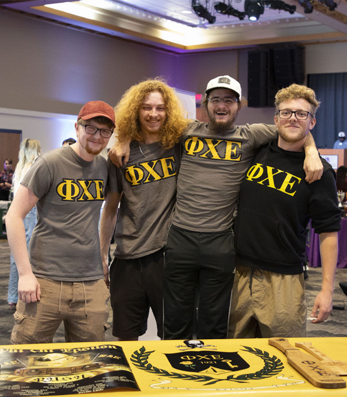 Four people wearing gray and yellow Phi Chi Epsilon shirts stand with their arms around each other's shoulders.