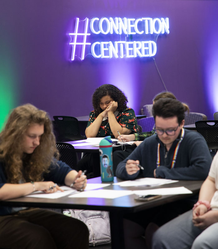 Three people sit at tables against a purple wall and a neon sign that says #connection centered.