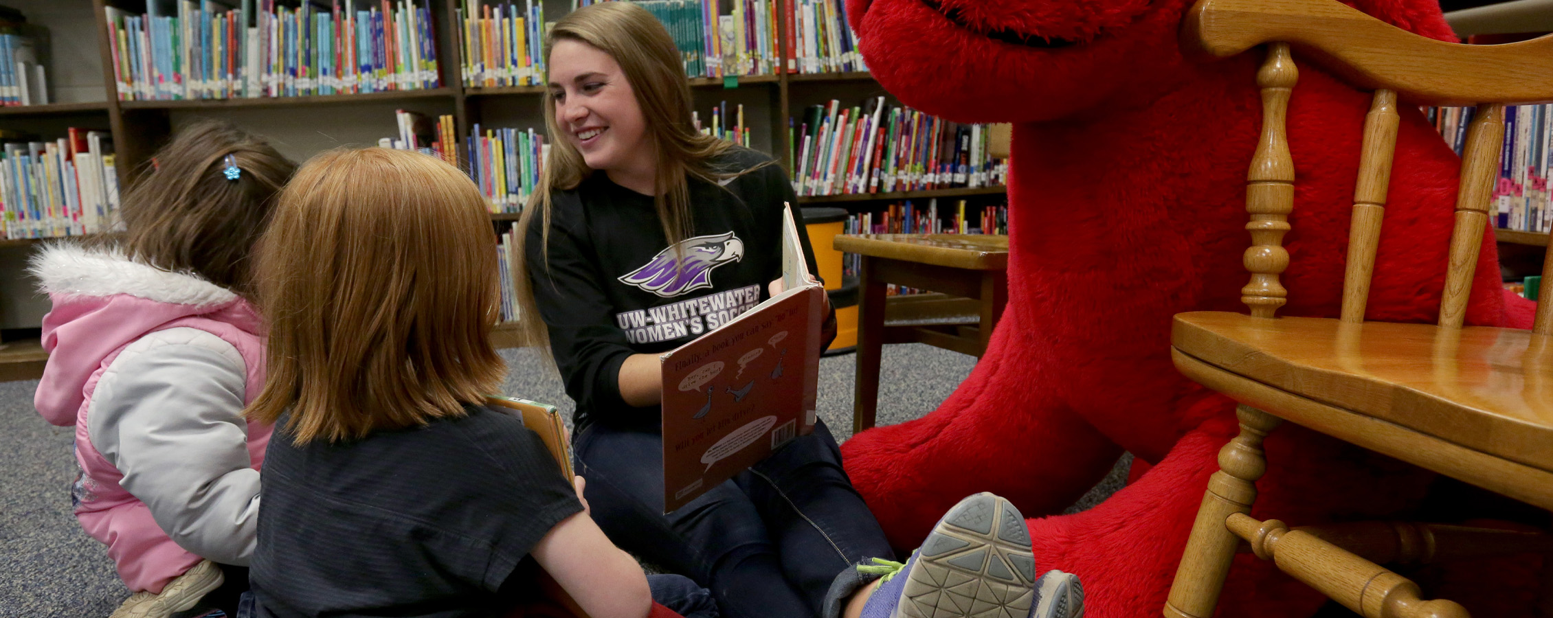 A Warhawk women's soccer player sits on the floor and reads with kids.