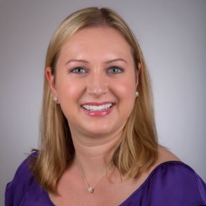 Deanna Guthrie smiles wearing a purple shirt in front of a grey background.
