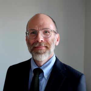 Edward Gimbel wearing glasses, a blue shirt and tie, and a blue suit jacket stands in front of a white wall with window light to his left.