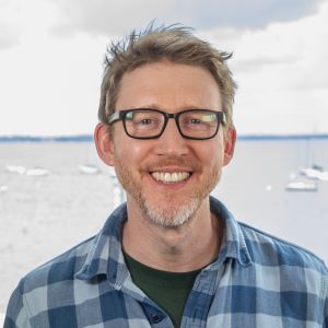 Headshot of Steven Girard outside wearing glasses, and a plaid shirt standing in front of water with boats in the background