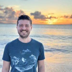 Alaz Kilicaslan stands on a beach wearing a blue t-shirt in front of water with a sunset in the background.