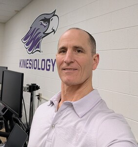 Mike Stibor stands in front of a white brick wall with a UW-Whitewater Warhawk and the word Kinesiology in the background.