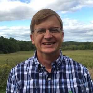 Rowand Robinson standing in a field with a blue sky behind him and wearing a blue plaid shirt.