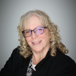 Sharri VanAlstine with glasses and curly blonde hair wears a black jacket sitting in front of a white wall.