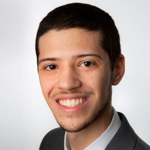 Chris Skrandzius smiles in front of a white wall while wearing a suit coat over a white shirt.