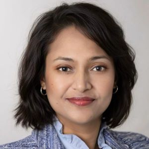 Headshot of Rimi Zakaria wearing a blue shirt and jacket standing in front of a white wall