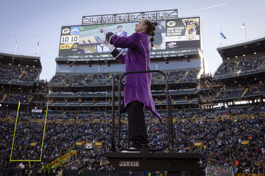 A student stand on an elevated platform and conducts with a Lambeau Field sign behind them.