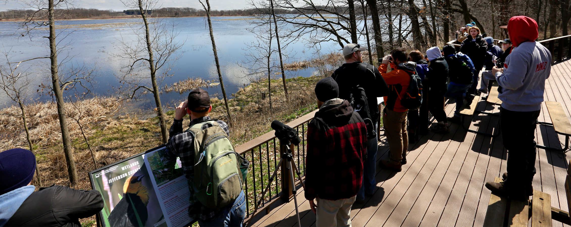 Students stand on a deck and look over Rose Lake State Natural Area through binoculars.