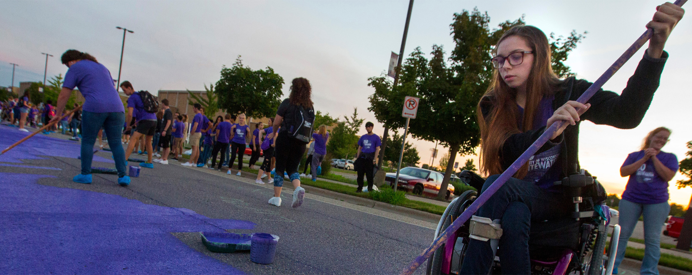 A student paints Warhawk Drive purple at the sun sets behind them.