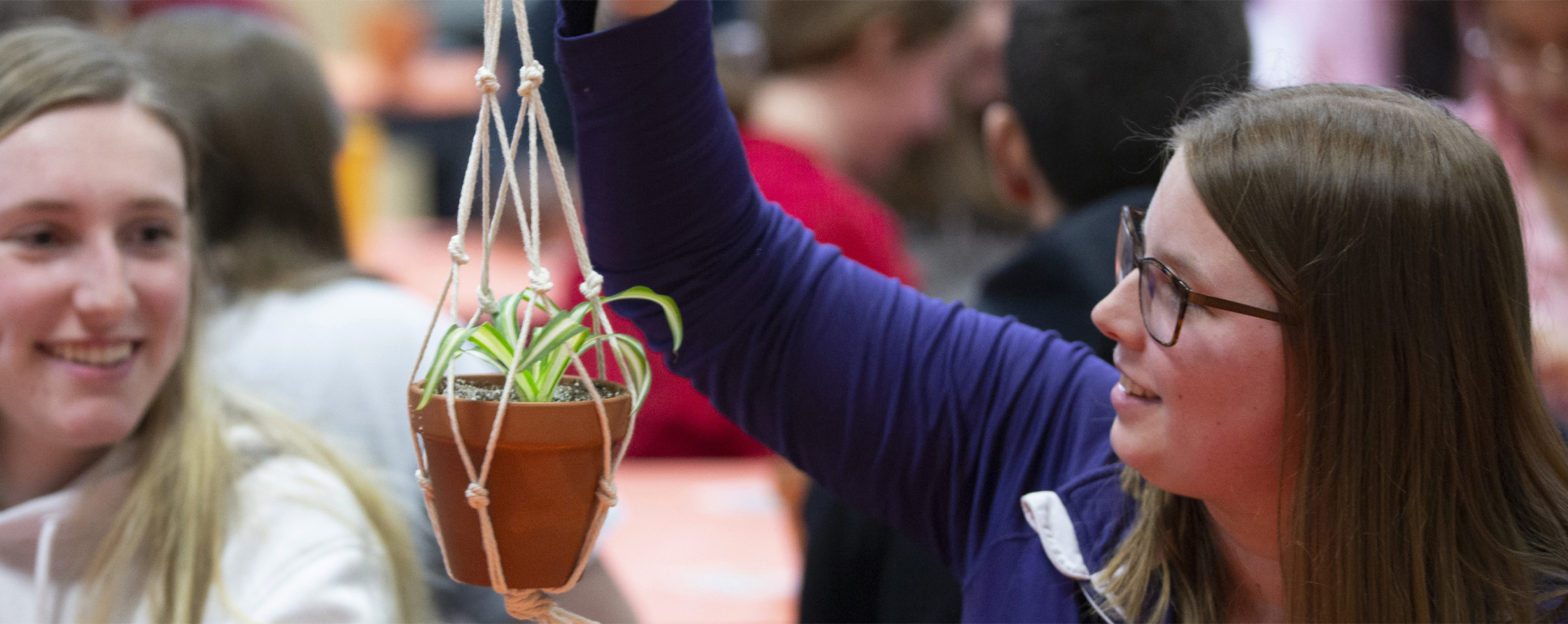 A student smiles and holds up a plant in a small macramé holder.