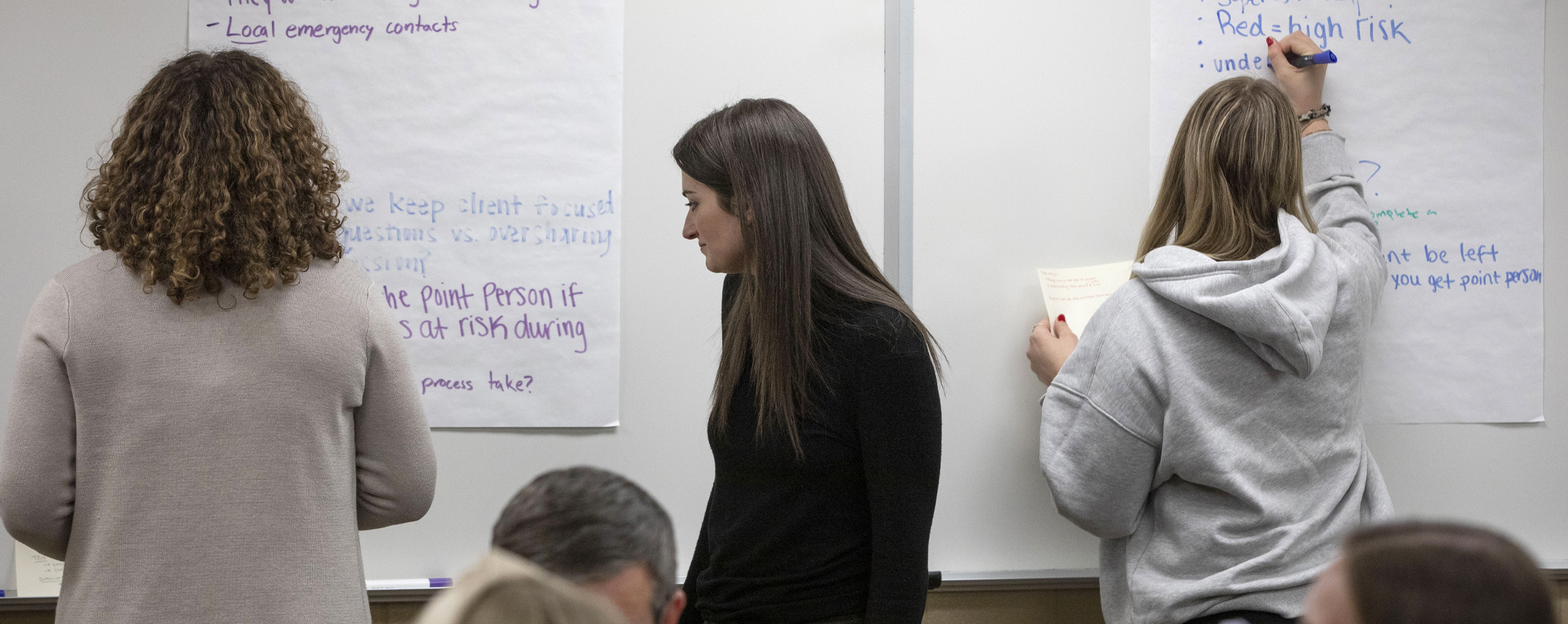 A faculty member stands with students who are writing on a board during an exercise in a classroom.