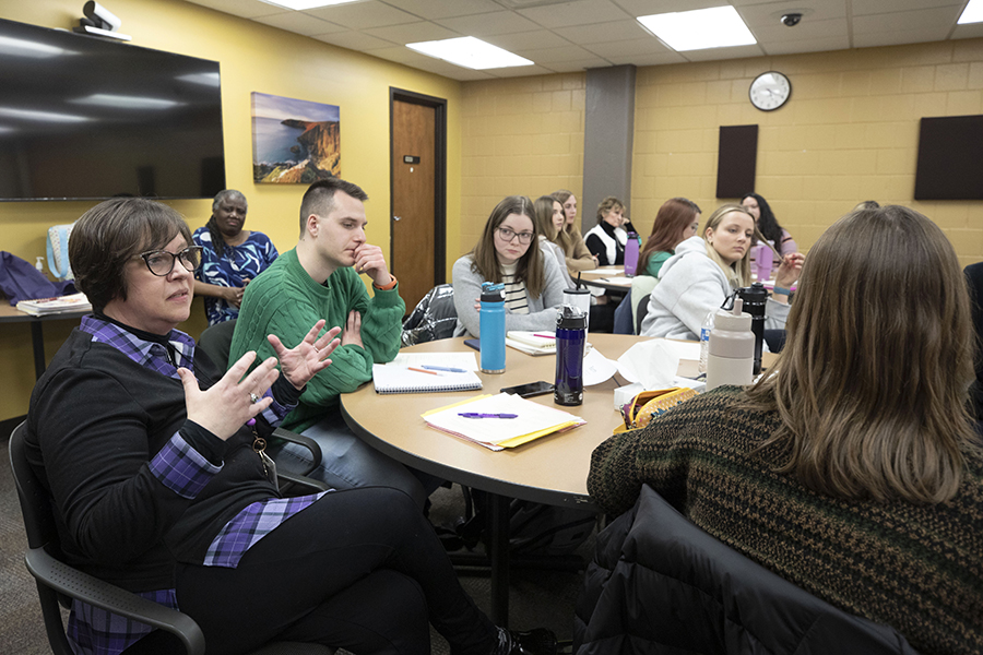 A faculty member sits and converses with students at a table.