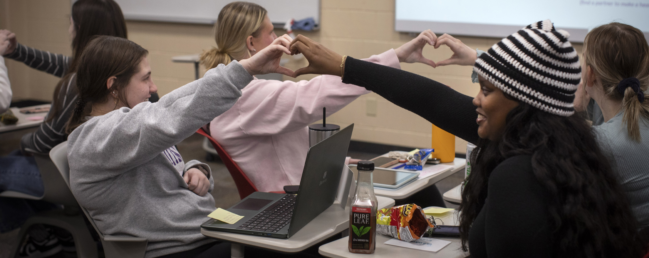 Students in a classroom sit across from each other and put their hands together to make hearts.