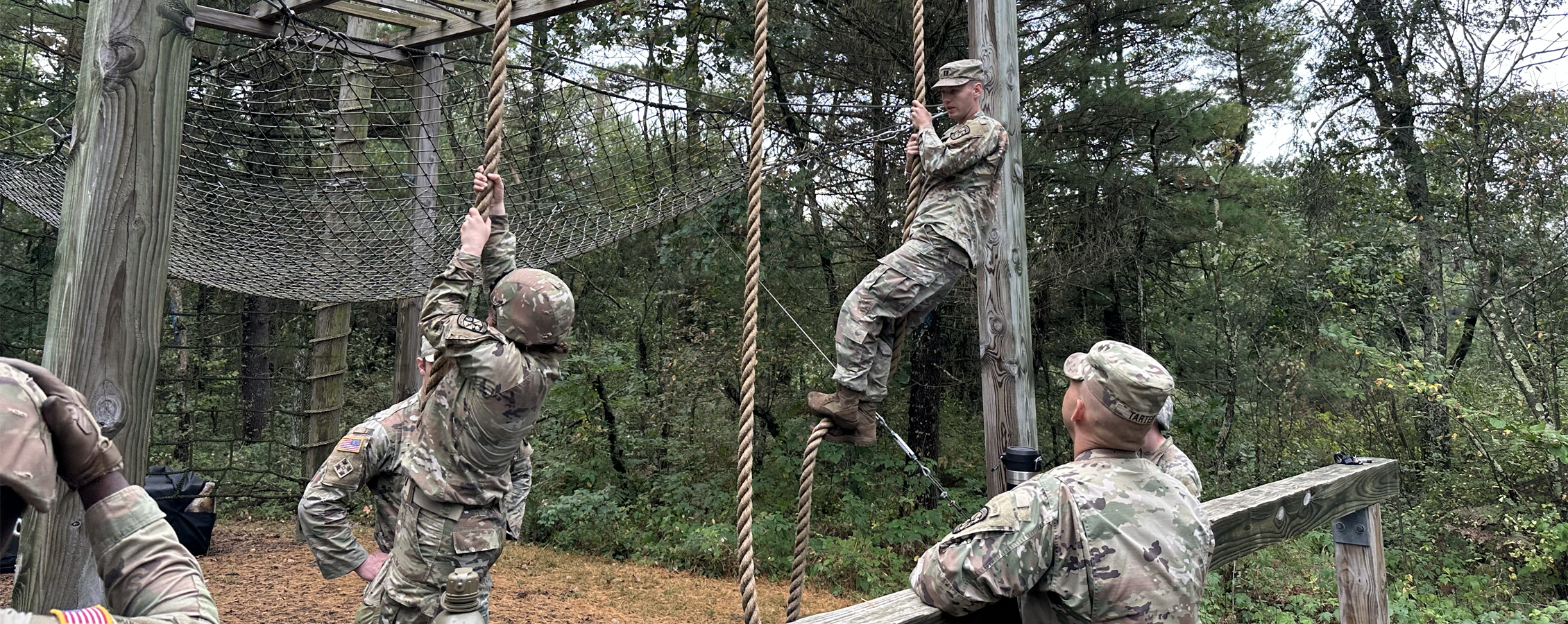 People in camo fatigues climb down ropes as part of an outdoor ropes course.
