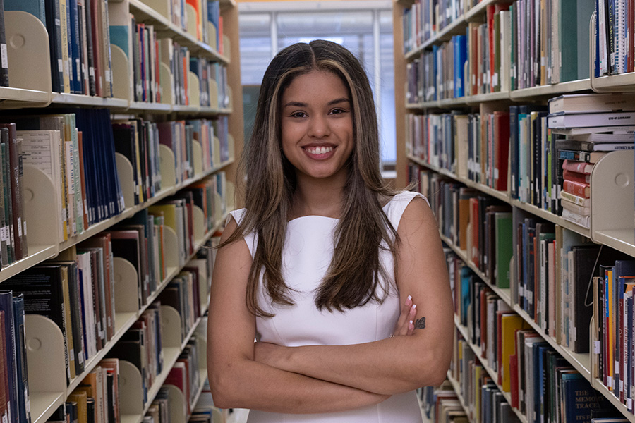 Kiara Rios wearing a white dress standing in between two library shelves with her arms crossed and smiling.