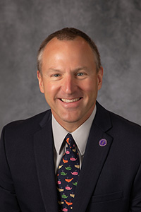 Pieter deHart wears a dark suit, white shirt, and colorful tie while sitting in front of a grey background.