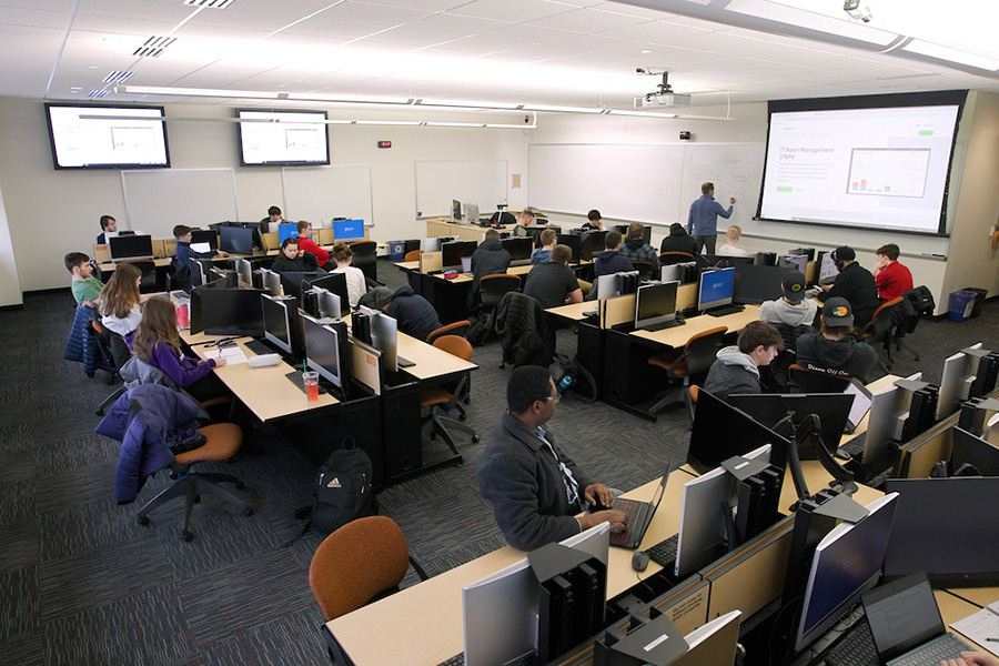 Classroom scene with students at computers and an instructor standing at a projector screen teaching.