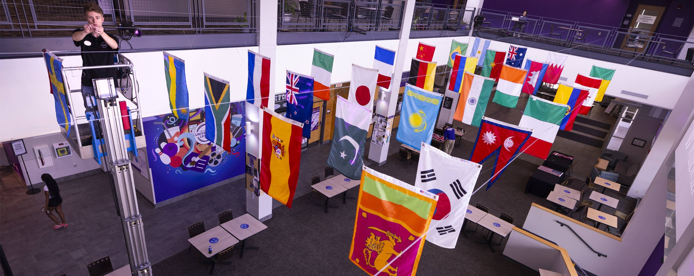 A person installs national flags above the commons area in a campus building.