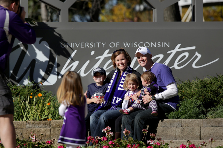 A family of two parents with children on their laps pose for a photo in front of a UW-Whitewater sign.