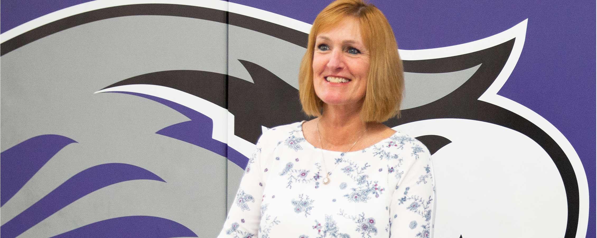 Kathleen Happel stands in front of a mat wall with a large Warhawk on it.