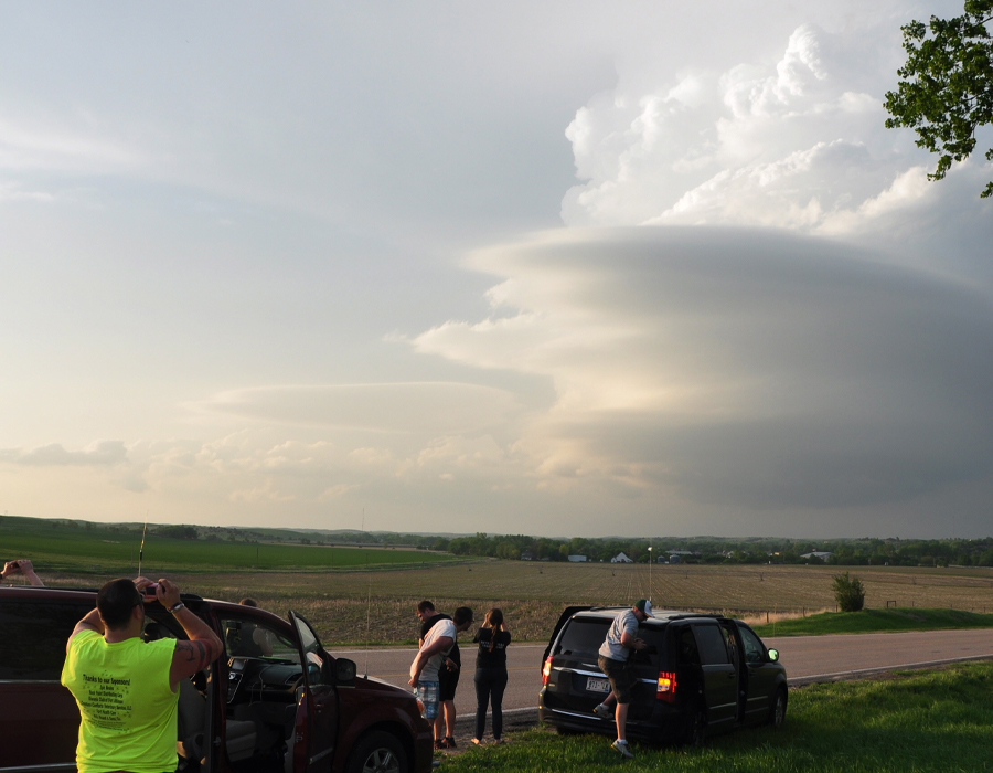 Students stand outside by vehicles, taking pictures of a stormy sky.