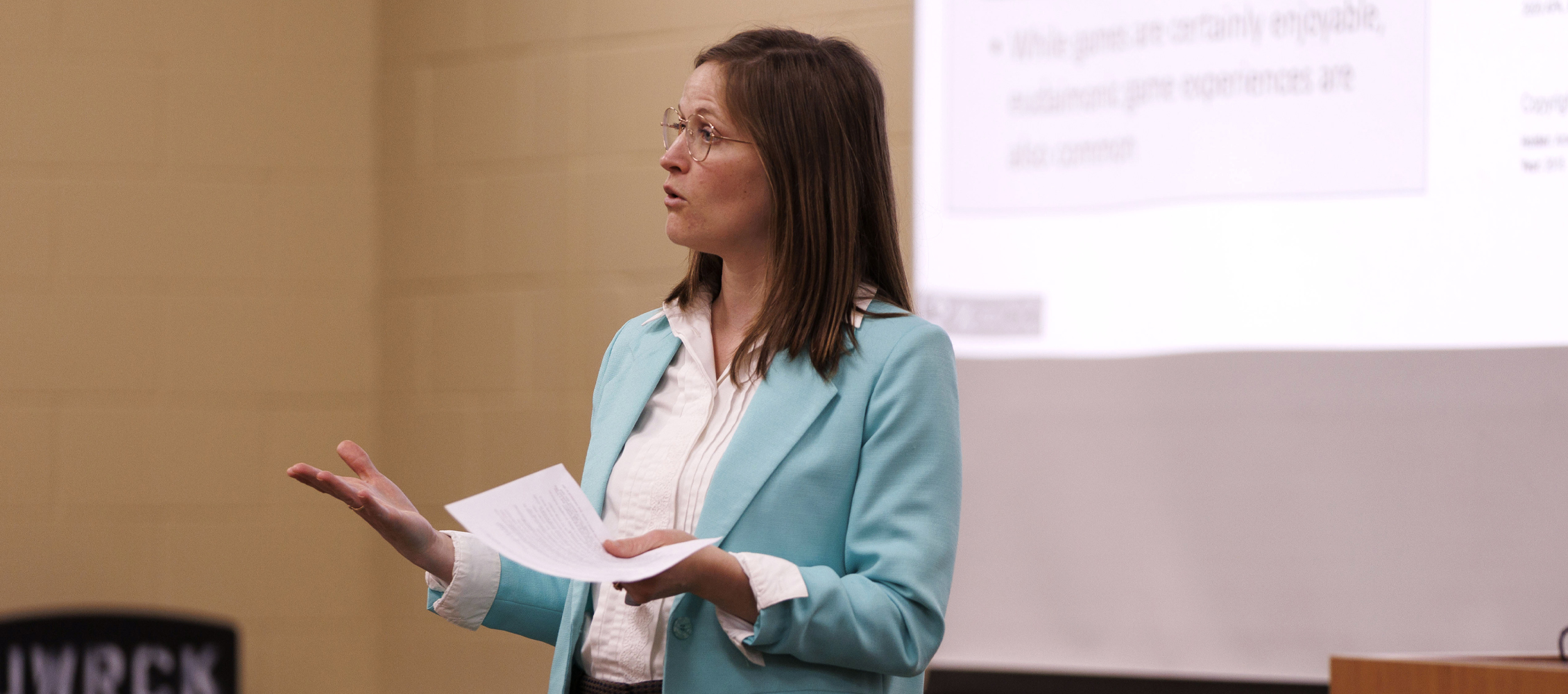 Kelly Jensen, wearing a teal blazer, uses her hands as she speaks at the front of class while holding a piece of paper.