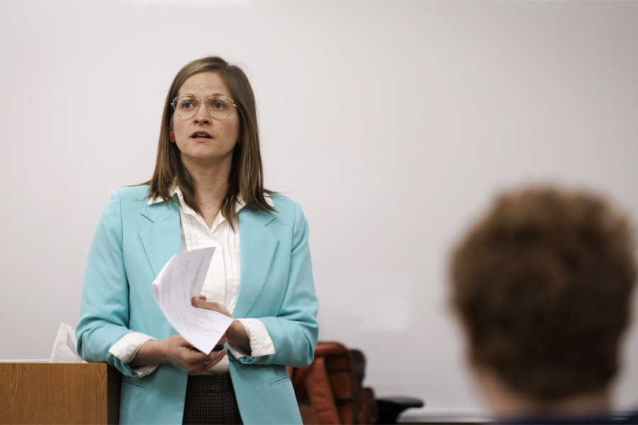 Kelly Jensen, wearing a teal blazer, speaks at the front of class while holding a piece of paper.