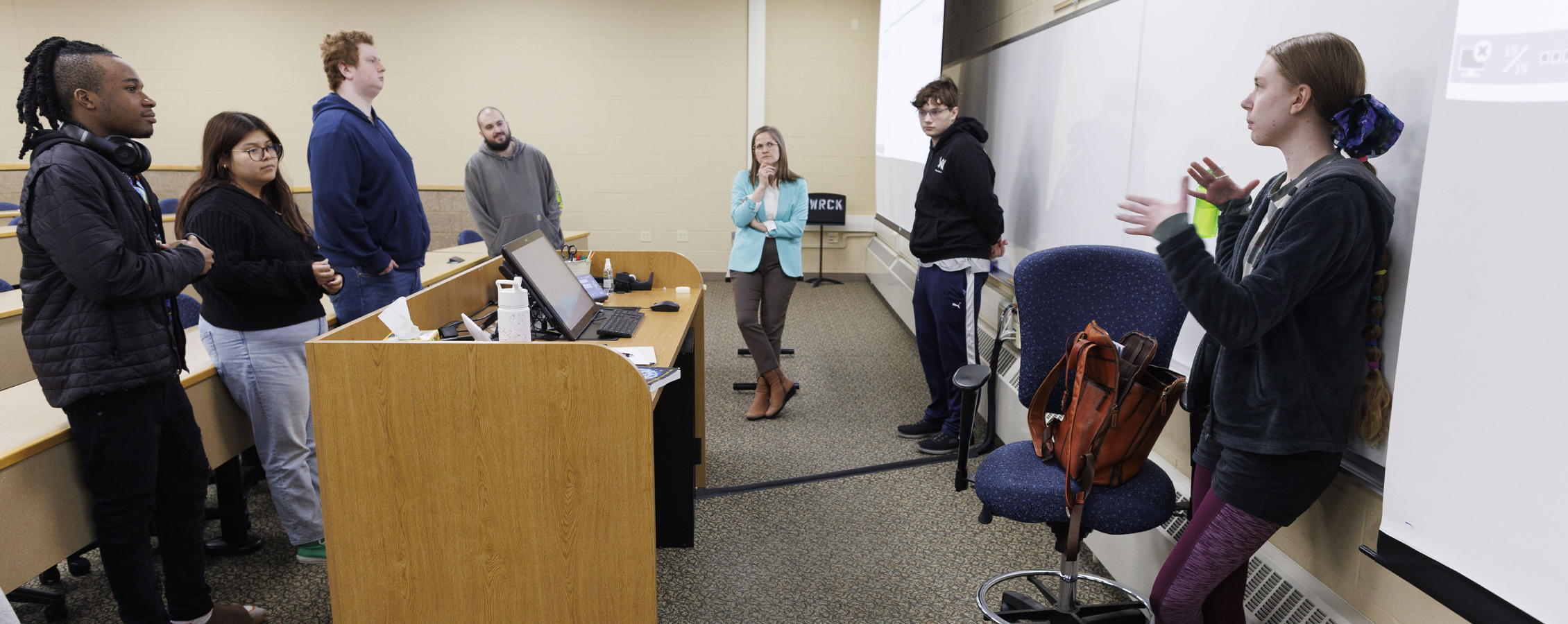 A group of students stand together and converse while Kelly Jensen looks on.