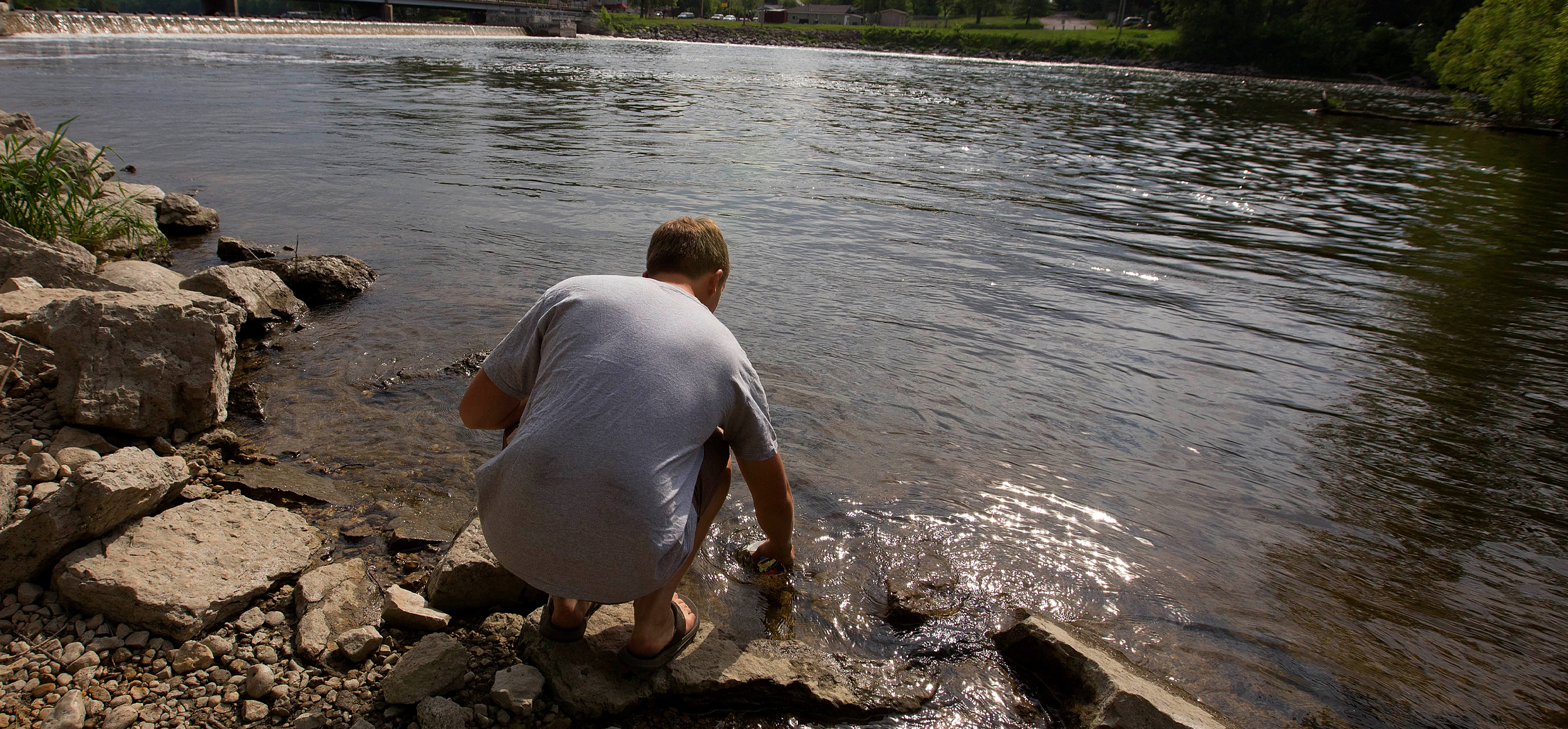 A person crouches on the edge of a river to collect a sample.