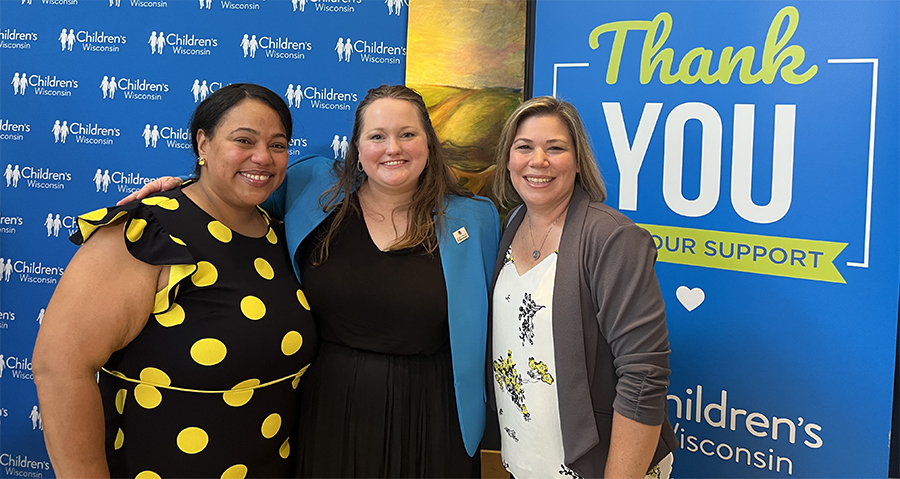 Three women stand together, smiling for the camera, in front of a blue Children's Hospital backdrop.