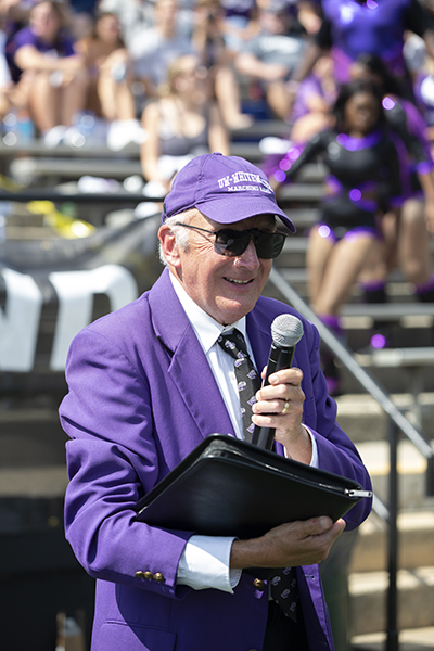 Glenn Hayes stands in Perkins Stadium with a microphone in his hand wearing a purple blazer.