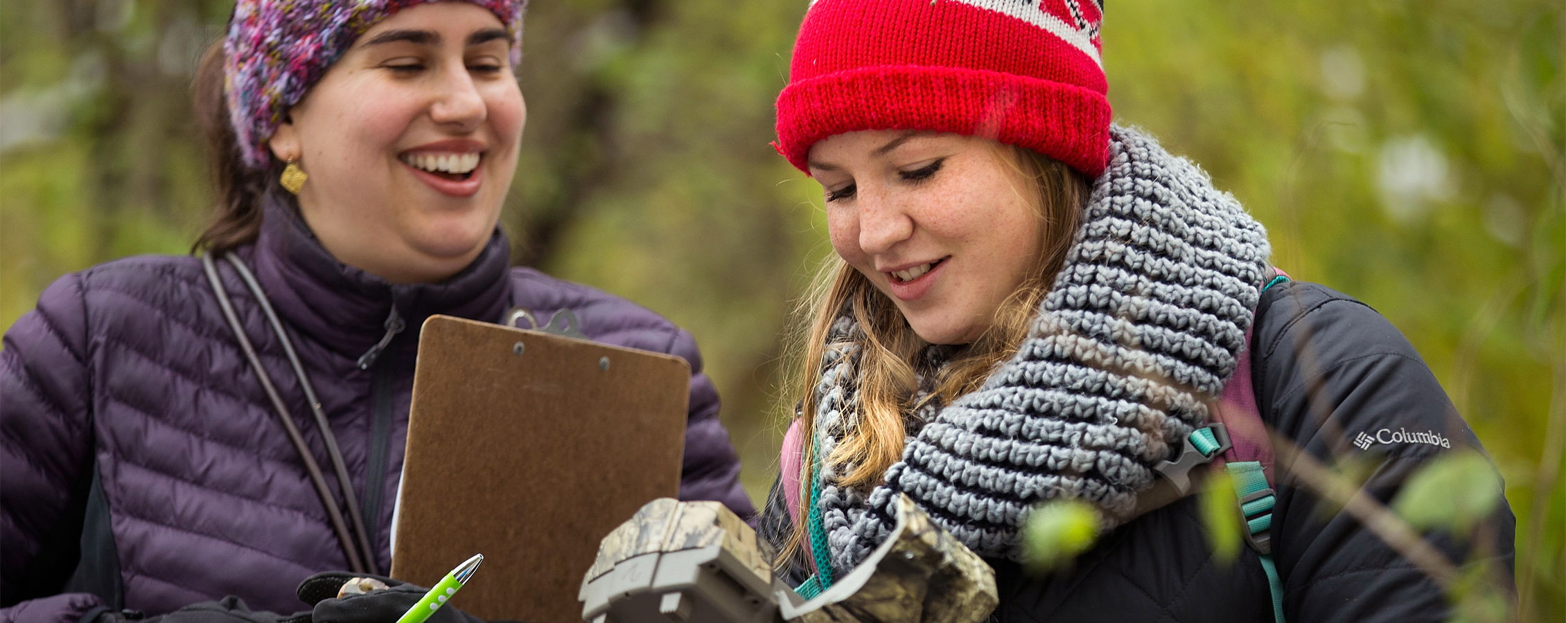 Two women are outside looking at a motion detecting trail camera.