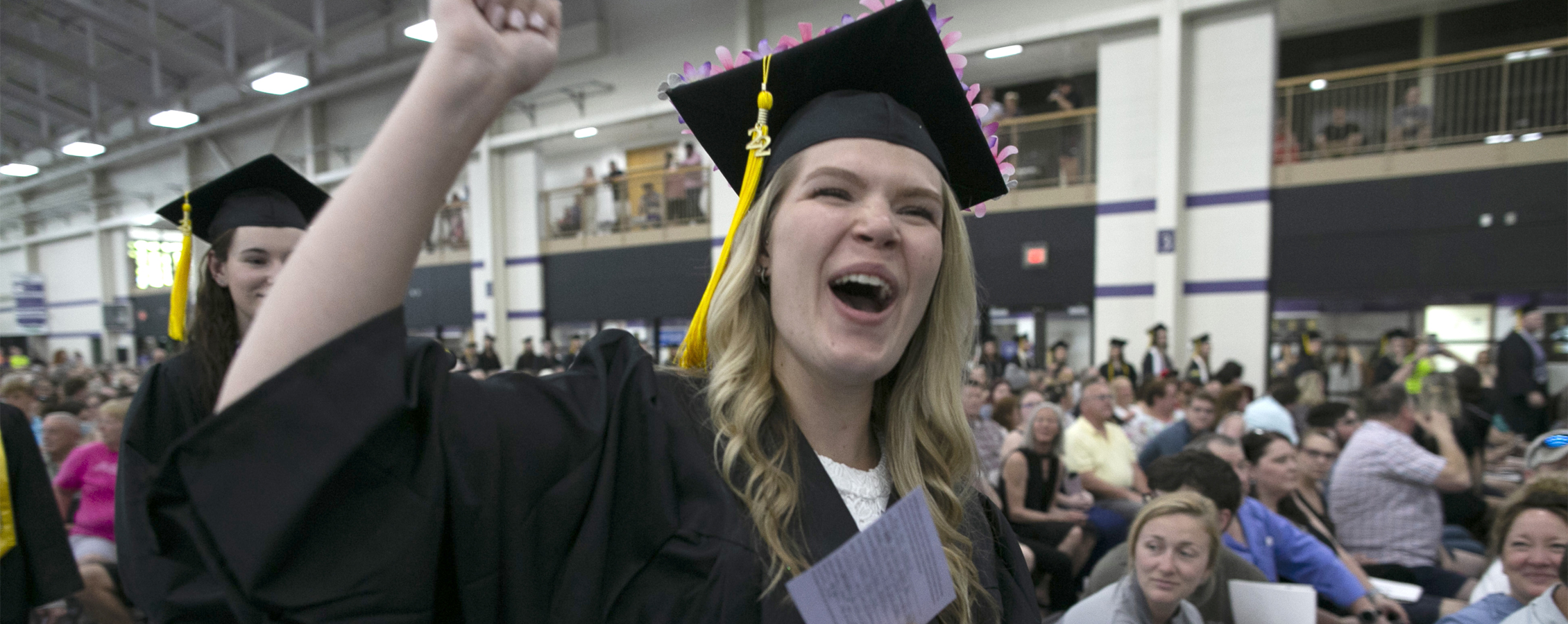 A student wearing her cap and gown cheers in the fieldhouse.