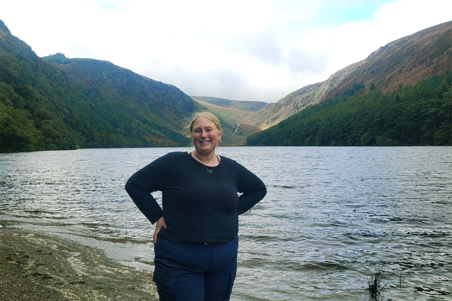 A person stands by the water in Ireland with mountains and sky in the background.