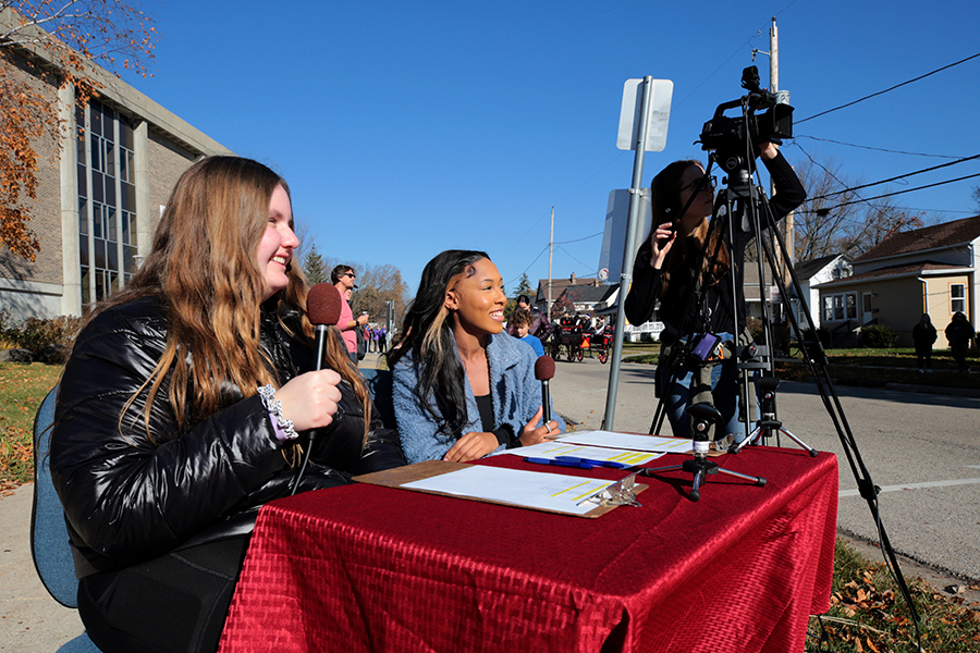 Carchesi sits at a table outside holding a microphone as the parade goes by.