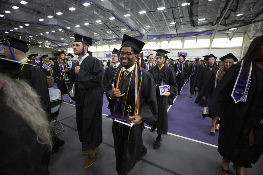 Graduates wearing cap and down walk down an isle in the fieldhouse during Commencement.