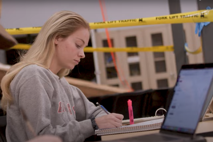 A student writes in a notebook while sitting in class with caution tape behind her.