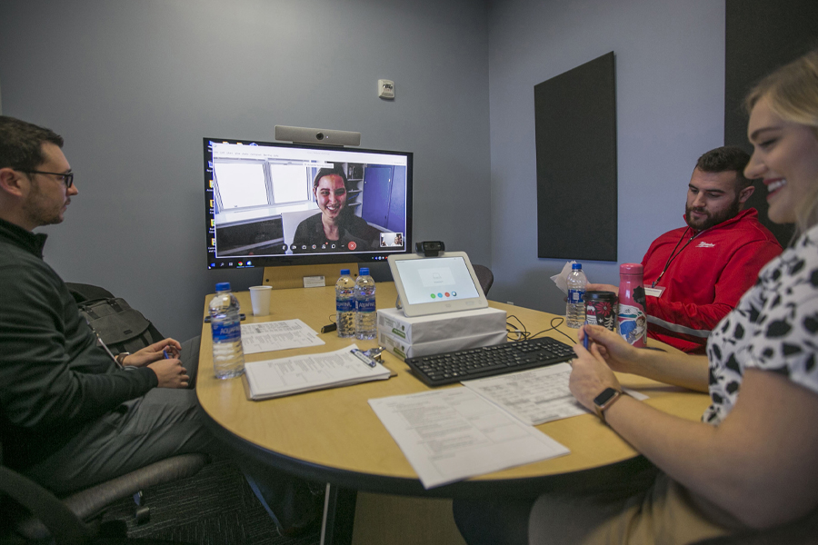Three people sit around a table with a monitor during a video conference.