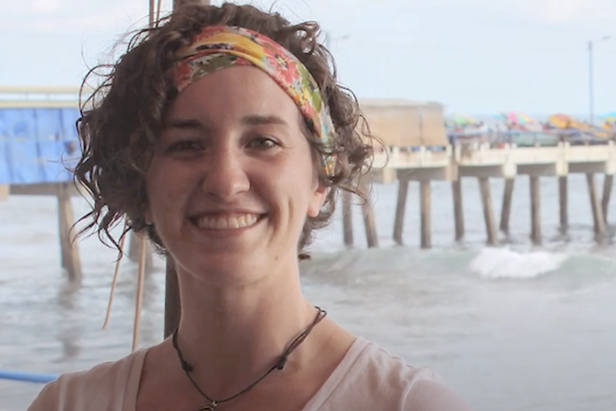 A person smiles at the camera with the sea in the background.