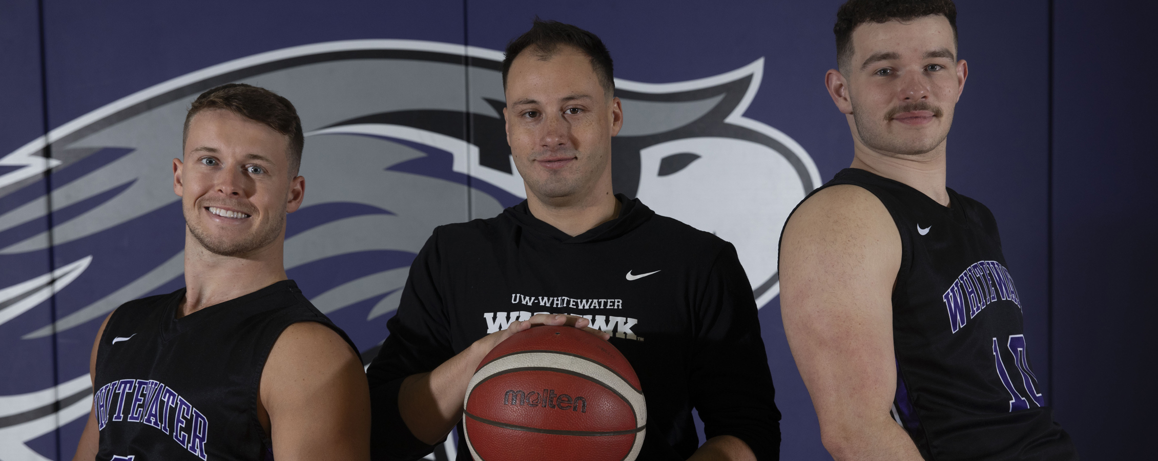Three Warhawks pose in front of a Warhawk wall and hold a basketball.
